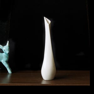 May include: A tall, elegant white ceramic vase with a slender, elongated body and a flared, asymmetrical opening. The vase has a smooth, glossy finish and stands on a dark wooden surface. A turquoise deer figurine is visible on the left.