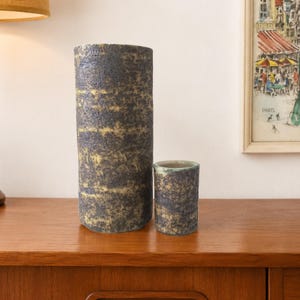 May include: Two cylindrical ceramic vessels with a rough, textured surface featuring a mottled dark charcoal and muted yellow finish. The taller vessel stands significantly higher than the shorter, wider-mouthed companion piece.
