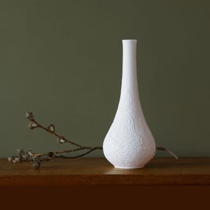 May include: A white ceramic vase with a textured, topographic pattern. The vase has a slender neck and a bulbous body. A dried branch with small seed pods rests on a wooden surface next to the vase.