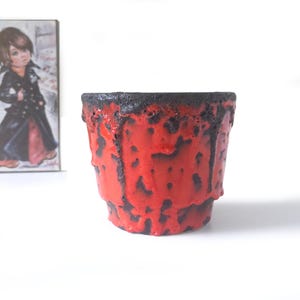 Può includere: Un vaso di ceramica rosso con un effetto smaltato strutturato e gocciolante. Il bordo superiore è nero, in contrasto con il corpo rosso vibrante. Il vaso è un piccolo oggetto decorativo, adatto per uso interno. Un'immagine incorniciata è sullo sfondo.