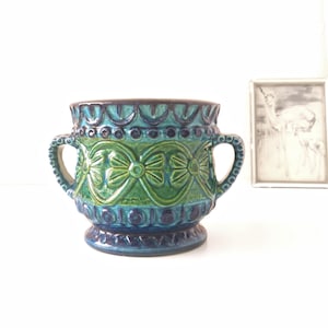 Pode incluir: Um vaso de cerâmica vintage com duas alças, apresentando um padrão detalhado em tons de verde-azulado, verde e azul. O vaso tem uma forma arredondada com uma base de pedestal e uma borda marrom escura. O design inclui motivos de laço repetidos.