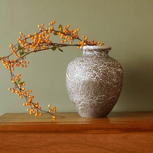 Könnte beinhalten: Eine dekorative Vase mit braun-weißem Craquelé-Finish. Die Vase enthält einen Zweig mit orangefarbenen Beeren und grünen Blättern. Die Vase steht auf einer Holzoberfläche.
