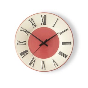 May include: A round wall clock featuring a cream-coloured outer ring with black Roman numerals and a solid coral-pink centre circle. The clock has three thin black hands positioned at the centre, with a thin coral-pink border around the outer edge.