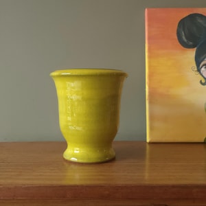 Puede incluir: Un jarrón de cerámica amarillo brillante con un borde acampanado y una base redondeada. El jarrón está sobre una superficie de madera. El fondo incluye una pintura con la cara de una mujer.