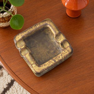 May include: A square, heavy-duty metal ashtray with a distressed, mottled gold and dark brown finish. The piece features four rounded notches along its thick, textured rim, designed for resting items, and a deep, smooth, dark central basin.