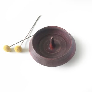 May include: A dark maroon ceramic ring holder with a central cone-shaped post, accompanied by two dried yellow billy ball flowers on thin stems. The bowl-shaped base has a smooth, matte finish and a slightly raised rim, providing a stable structure.