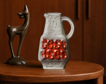 May include: A ceramic pitcher with a textured white glaze and a prominent orange relief pattern on the front, featuring a single handle and a small spout. Beside it stands a smooth, dark brown ceramic figurine of a stylized deer.