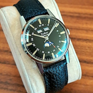 May include: A vintage Zodiac automatic watch with a black dial and a black leather band. The watch face displays the day, date, and moon phase. The watch is set on a beige cushion, showcasing its classic design and craftsmanship.
