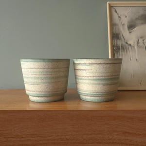 Puede incluir: Dos macetas de cerámica con base moteada color crema y rayas horizontales en verde y morado apagados. Las macetas tienen una forma ligeramente acampanada y están sobre una superficie de madera.