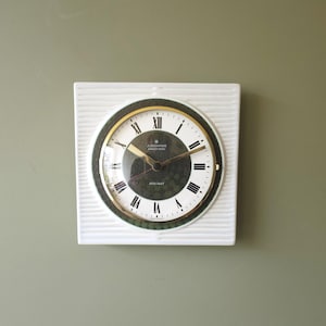 Può includere: Un orologio da parete quadrato bianco con un quadrante strutturato. Il quadrante è rotondo con una lunetta dorata, numeri romani neri e lancette dorate. L'orologio indica "Junghans Electronic ATO-MAT".