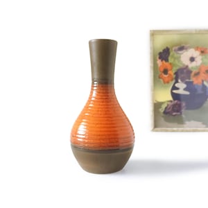 Può includere: Un vaso in ceramica con collo e base marroni e corpo arancione strutturato. Il vaso ha una forma arrotondata e un collo stretto. Un'immagine incorniciata con fiori è sullo sfondo.