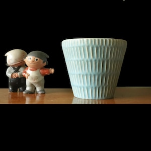 Puede incluir: Un jarrón de cerámica azul claro con un diseño acanalado vertical texturizado. Dos pequeñas figuras vintage están a la izquierda del jarrón. Las figuras visten ropa vintage. El fondo es negro.