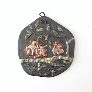 Peut inclure: Plaque murale en céramique représentant trois oiseaux perchés sur une branche. Les oiseaux sont rouges et bruns avec des détails jaunes. Le fond est gris foncé avec un motif d'arbre et de feuilles. Une boucle noire est en haut pour accrocher.