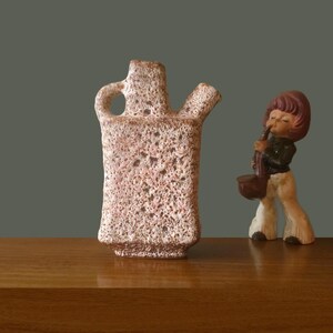 Peut inclure: Un vase en céramique texturé et clair avec une poignée et deux becs se dresse sur une surface en bois. À côté, une figurine d'un enfant jouant du saxophone. La figurine porte une veste noire et un pantalon blanc.