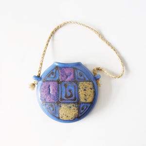May include: A round blue ceramic wall pocket featuring a grid of square panels with purple and yellow speckled glazes and dark brown spiral motifs. The vessel includes two side loops attached to a braided tan rope for hanging on a wall surface.