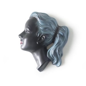Puede incluir: Una escultura de pared de cerámica que muestra el perfil de un rostro con piel negra mate, labios rojos y cabello ondulado de color azul claro recogido en una coleta. La pieza presenta contornos suaves y un acabado brillante en el pelo.