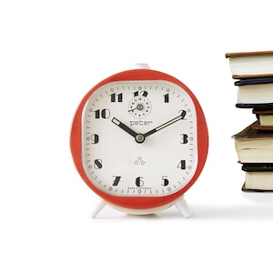 Puede incluir: Un reloj despertador retro rojo y blanco con el nombre "peter" en la esfera. El reloj tiene una esfera blanca con números y manecillas negras. Una pila de libros es visible en el fondo.