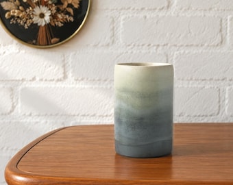 May include: A cylindrical ceramic vase featuring a smooth gradient finish that transitions from a pale cream colour at the top to a deep, muted charcoal grey at the base. The surface has a matte texture with subtle, horizontal colour blending.