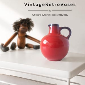 May include: A glossy red ceramic pitcher with a rounded body, a small spout, and a dark blue handle. The text VintageRetroVases and AUTHENTIC EUROPEAN DESIGN 1950s-1980s is visible at the top. A wooden monkey figurine is positioned in the background.
