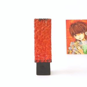 May include: A tall, rectangular ceramic vase with a textured, orange glaze and a black base. The vase is standing on a white surface.