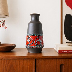 May include: A tall ceramic vase with a matte dark brown finish, featuring a wide central band of vibrant orange glaze decorated with raised, swirling teal blue patterns and a geometric border design around the top and bottom of the band.