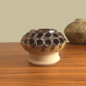Puede incluir: Un pequeño jarrón de cerámica con un diseño único. El jarrón presenta un cuerpo marrón texturizado con múltiples aberturas circulares alrededor de la parte superior. La base es de color beige claro. Otro jarrón está en el fondo.