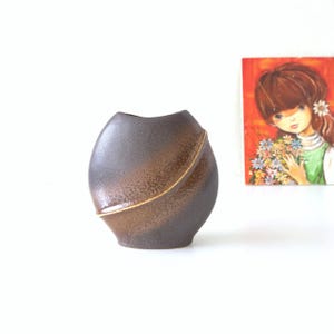 May include: A brown ceramic vase with an unusual, asymmetrical form. The vase has a dark brown base that fades to a lighter brown with a gold accent line. The vase is approximately 18 cm tall.