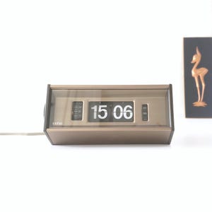 May include: A vintage Copal flip clock with a gold-tone case and a clear plastic cover. The clock displays the time as 3:06 PM in large, white numerals. The clock has a retro design, with a small dial on the left side and a minute dial on the right.