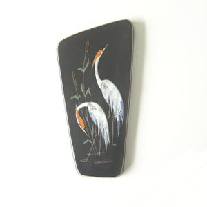 May include: A black wall art piece with a silver trim, featuring a painted scene of two white herons with orange accents and blue wing tips, standing in water with cattails. The artwork has a unique, asymmetrical shape.