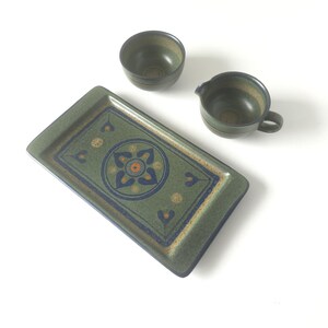 May include: A ceramic serving set featuring a rectangular tray, a small bowl, and a creamer. The set has a green base color with blue, orange, and yellow geometric patterns. The tray measures approximately 12 inches long.