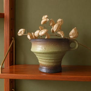 May include: A ceramic vessel with a wide, flared rim and a single side handle, featuring a textured, gradient glaze that transitions from dark brown at the base and rim to a muted sage green in the centre, containing dried, papery seed pods.
