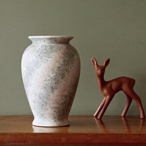 Puede incluir: Un jarrón de cerámica moteado, de color blanco roto, con una base ancha y un cuello estrecho, se encuentra junto a una pequeña figura de ciervo marrón. El jarrón tiene un aspecto texturizado con acentos rosas y grises. El ciervo está posando con las patas extendidas.