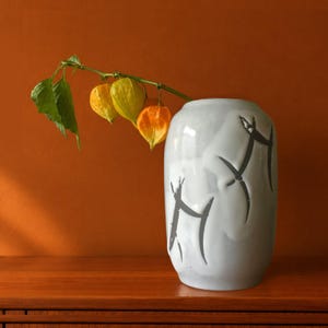 Op de afbeelding: Een lichtgrijze keramische vaas met een glad, afgerond silhouet voorzien van donkere, abstracte, penseelstreekachtige markeringen. Een enkele groene stengel steekt uit de bovenkant, met drie levendige oranje, lampionvormige physalisvruchten.