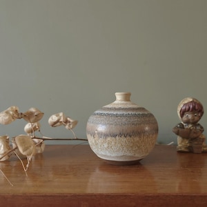 Puede incluir: Un jarrón de cerámica redondo con un patrón de rayas beige y marrón. El jarrón tiene un cuello pequeño y cilíndrico y está sobre una superficie de madera. También se ve una pequeña figura y tallos de plantas secas.