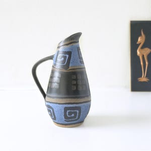 Könnte beinhalten: Keramikkrug mit schwarzem Griff und gebogenem Ausguss. Der Krug hat ein Design aus blauen und schwarzen geometrischen Formen mit einem beigefarbenen Band in der Nähe des Bodens. Der Krug ist wahrscheinlich Vintage oder handgefertigt.