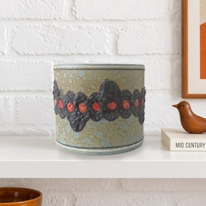 Può includere: Un vaso cilindrico in ceramica con una smaltatura di base maculata verde chiaro e blu. Un'ampia fascia testurizzata grigio antracite avvolge il centro, punteggiata da una fila orizzontale di piccoli accenti decorativi in rilievo rosso-arancio.