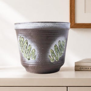 Pode incluir: Um vaso de cerâmica castanho-escuro com uma borda larga e alargada e uma base cónica. O exterior apresenta um vidrado branco difuso no topo e dois padrões verticais em ziguezague de cor verde-claro texturizados no centro do corpo.