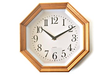 Puede incluir: Reloj de pared octogonal con marco de madera marrón claro. La esfera del reloj es blanca con números y manecillas negras. La palabra "QUARTZ" está impresa en la parte inferior de la esfera.