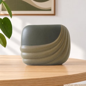 Può includere: Un vaso in ceramica arrotondato con un design bicolore, caratterizzato da una sezione superiore grigio antracite scuro e una sezione inferiore verde salvia testurizzata. Il vaso presenta una serie di scanalature orizzontali curve sul lato destro.