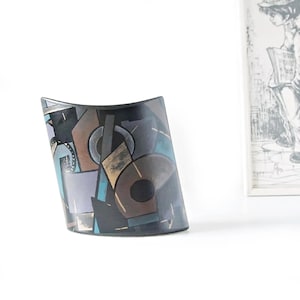 May include: A black, curved vase with an abstract geometric design. The design features shapes in shades of brown, blue, gray, and lavender. The vase has a glossy finish and is set against a white background.