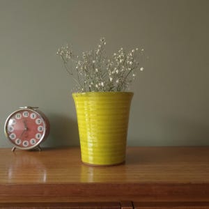 Puede incluir: Un jarrón de cerámica amarillo brillante con un diseño acanalado, lleno de delicadas flores blancas. Un reloj despertador vintage con esfera roja y números blancos está a la izquierda. Sobre una superficie de madera.