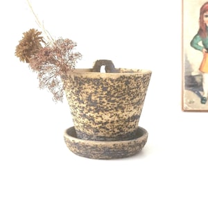 Op de afbeelding: Een gespikkelde keramische plantenbak met een bijpassende schotel. De plantenbak heeft een handvat en is gevuld met gedroogde bloemen. De plantenbak is ongeveer 10 cm hoog en 13 cm breed.