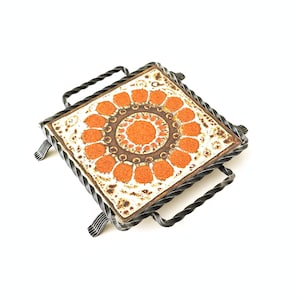 Puede incluir: Una baldosa cuadrada de cerámica con un diseño floral marrón y naranja está enmarcada en un marco de metal negro con asas de alambre retorcido. La baldosa es un salvamanteles o una almohadilla caliente.
