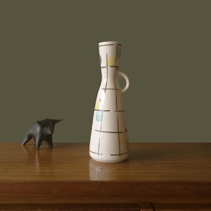 May include: A tall, cream-colored ceramic vase with a small handle and a geometric pattern of black lines and colorful squares. A small, dark bronze bull figurine stands nearby. The vase is approximately 10 inches tall.