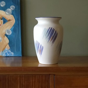 Puede incluir: Un jarrón de cerámica blanco con borde acampanado y diseño de pinceladas abstractas en tonos azules, morados y naranjas. El jarrón se exhibe sobre una superficie de madera.