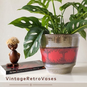 Op de afbeelding: Een keramische plantenpot met een glanzende grijze glazuurlaag aan de boven- en onderkant en een brede, getextureerde band van felrood in het midden. De rand heeft een decoratief ingesprongen patroon. Een groene plant met geperforeerde bladeren staat erin.