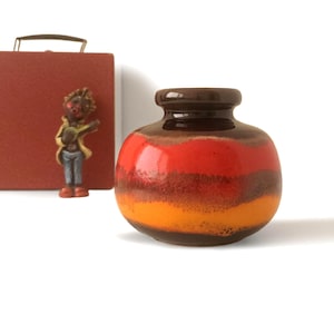 Può includere: Un piccolo vaso rotondo in ceramica con parte superiore e base marrone scuro e un design a strati di rosso, marrone e arancione. Una miniatura di una persona che suona la chitarra e una valigetta rossa sullo sfondo.