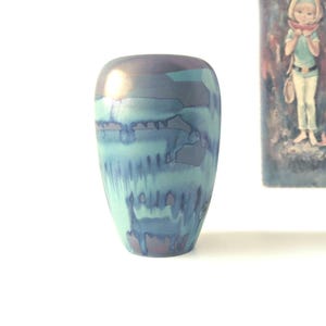May include: A tall, ceramic vase with a rounded top and a flared base. The vase features a blend of blue, teal, and purple hues with a glossy finish. The design has a watercolor effect, with streaks and patches of color. The vase is a decorative item.