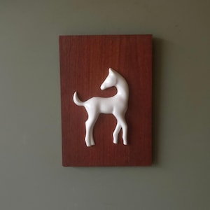 May include: A rectangular wall plaque featuring a smooth, white, three-dimensional relief of a standing foal centered on a dark, reddish-brown wooden board. The foal is depicted in profile, facing left, with a raised tail and slender legs.