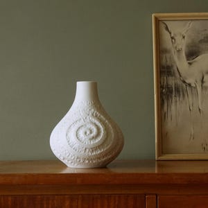 Può includere: Un vaso in ceramica bianca con finitura opaca e corpo arrotondato. La superficie presenta una prominente trama a spirale in rilievo che ricorda il motivo di una conchiglia fossilizzata, estendendosi dal centro verso l'esterno.